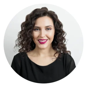 Portrait d’une femme aux cheveux bouclés, sourire, rouge à lèvres rose, haut noir, fond clair.